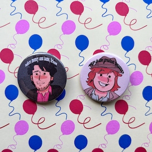 May include: Two circular pin-back buttons featuring illustrated characters. The left button shows a person with dark hair and the text "before money and fame, friend". The right button shows a person in a hat with the text "ship is the greatest treasure".