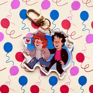 Puede incluir: Un llavero de acrílico con una ilustración de dibujos animados de dos personajes con mejillas sonrojadas. Un personaje tiene el pelo rojo y un sombrero marrón, mientras que el otro tiene el pelo oscuro. Se ven un corazón y una nota musical.