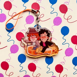May include: An acrylic charm featuring two illustrated characters riding a large orange bottle with the text "orbit" printed on it. One character wears a brown hat and coat, while the other has dark hair and a black suit. It includes a metal clasp and orange cord.