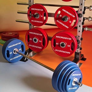 Peut inclure: Un ensemble d'haltérophilie avec une barre et des disques bleus, ainsi qu'un rack de disques rouges. Les disques affichent la marque "MICROGYM" et des indications de poids. Le fond est un dégradé orange et gris. Les poids sont en kilogrammes.