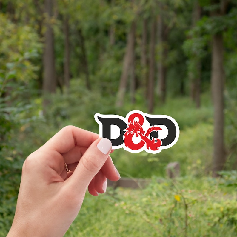 Może przedstawiać: Naklejka z literami "D&D" w kolorze czarnym i czerwonym, ze wzorem smoka. Naklejka jest prezentowana na rozmytym zielonym tle drzew i trawy.