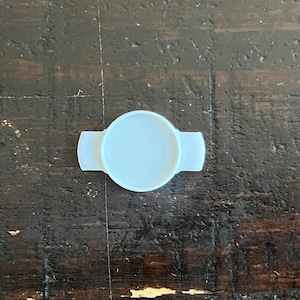 Può includere: Un tappo per bottiglia azzurro chiaro, di forma circolare, con due estensioni rettangolari su entrambi i lati. Il tappo è realizzato in un materiale liscio e solido e poggia su una superficie di legno marrone scuro strutturata. Il design è semplice e funzionale.