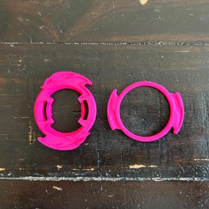 May include: Two bright pink, circular plastic components with a geometric design. One has a central opening and stylized edges, while the other is a simple ring with flared sections. They are displayed on a dark wooden surface.