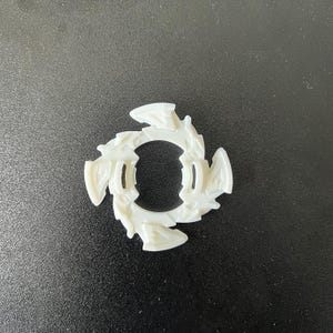 May include: A white plastic toy component with a circular design. The component features a series of stylized, wing-like shapes around its perimeter. The center has a circular opening. The toy part is designed for a children's game.