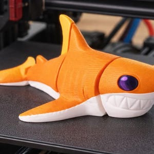 Pu&ograve; includere: Una statuetta di squalo arancione stampata in 3D con dettagli bianchi e un occhio viola. Lo squalo ha un corpo segmentato e poggia su una superficie nera. I denti dello squalo sono bianchi e le pinne gialle.