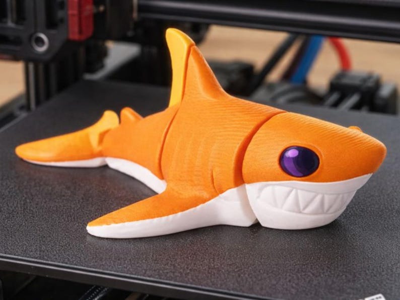 Pu&ograve; includere: Una statuetta di squalo arancione stampata in 3D con dettagli bianchi e un occhio viola. Lo squalo ha un corpo segmentato e poggia su una superficie nera. I denti dello squalo sono bianchi e le pinne gialle.