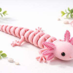 May include: A pink axolotl figurine with a segmented body and feathery gills. The salamander-like creature has small legs and a smiling face. The figurine is set against a white background with decorative elements.