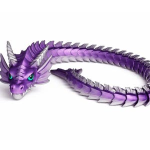 Pu&ograve; includere: Una statuetta di drago viola con un design dettagliato. Il drago ha una testa con corna, occhi verdi e un corpo lungo e articolato. Le scaglie sono una sfumatura di viola e argento.