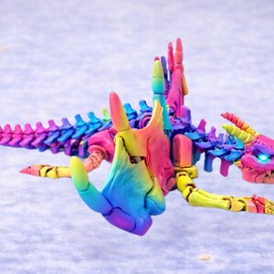 Op de afbeelding: Een veelkleurig, beweegbaar drakenbeeldje. De draak heeft een gedetailleerde skeletstructuur en een kop met oplichtende blauwe ogen. De kleuren zijn roze, geel, blauw en groen. Een opvallend en gedetailleerd fantasiefiguur.