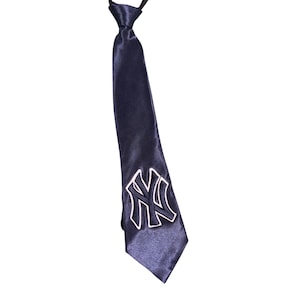 Könnte beinhalten: Eine marineblaue Krawatte mit einem weißen, gestickten "NY"-Logo am unteren Ende. Die Krawatte besteht aus einem glänzenden, satinartigen Material und hängt vor einem weißen Hintergrund.