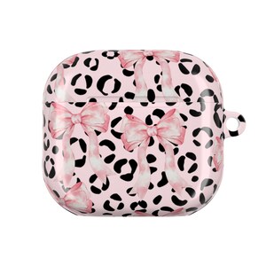 Könnte beinhalten: Ein rosa AirPod-Case mit einem sich wiederholenden Muster aus schwarzen Leopardenflecken und rosa Schleifen im Aquarellstil. Das Case hat ein glänzendes Finish und eine kleine Schlaufe zum Befestigen an einem Schlüsselanhänger. Das Design ist feminin und stilvoll.
