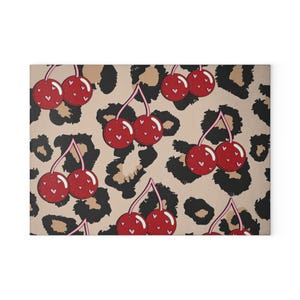 Könnte beinhalten: Ein rechteckiger dekorativer Teppich mit einem sich wiederholenden Muster aus roten Kirschen mit Herzdetails auf einem beigefarbenen Hintergrund mit schwarzen und braunen Leopardenflecken. Die Kirschen haben rosa Stiele.