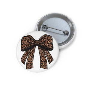 Könnte beinhalten: Ein weißer Button mit einem Leopardenmuster-Schleifendesign. Der Bogen hat braune und schwarze Flecken auf einem beigen Hintergrund. Ein zweiter Button ist im Hintergrund sichtbar.