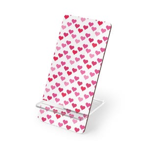 Könnte beinhalten: Ein weißer Handy-Ständer mit einem Muster aus roten und rosa Herzen. Der Ständer hat einen transparenten Kunststofffuß und einen schwarzen Rand. Die Herzen sind in verschiedenen Rosa- und Rottönen gehalten. Ein dekoratives Accessoire.