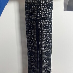 May include: A rectangular black crochet wall hanging featuring a central sword motif surrounded by floral patterns. The textile is suspended from a light-colored wooden dowel at the top and weighted by a matching wooden bar at the bottom edge.