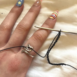 Könnte beinhalten: Ein silberfarbener Metallring in Form einer Katze mit langem Schwanz, der an einem Finger getragen wird. Ein dünner schwarzer Faden verläuft durch den katzenförmigen Ring und ist mit einer silbernen Häkelnadel verbunden, die eine Kette aus schwarzen Maschen bildet.