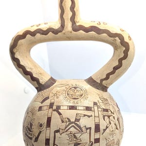 May include: A cream-colored ceramic stirrup-spout vessel featuring dark brown hand-painted illustrations. The spherical body displays intricate figures, including a central character in a frame, surrounded by geometric patterns and sun-like motifs.