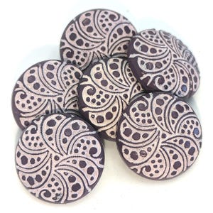 May include: Six round, dark purple buttons with a white, intricate, floral design. The buttons have a raised, textured surface.