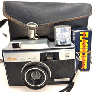 May include: A black and silver 35mm film camera with a flash attachment and a leather carrying case. The camera has the text "skan ELECTRO-MATIC" and "TEN FIFTY FOUR" printed on it.