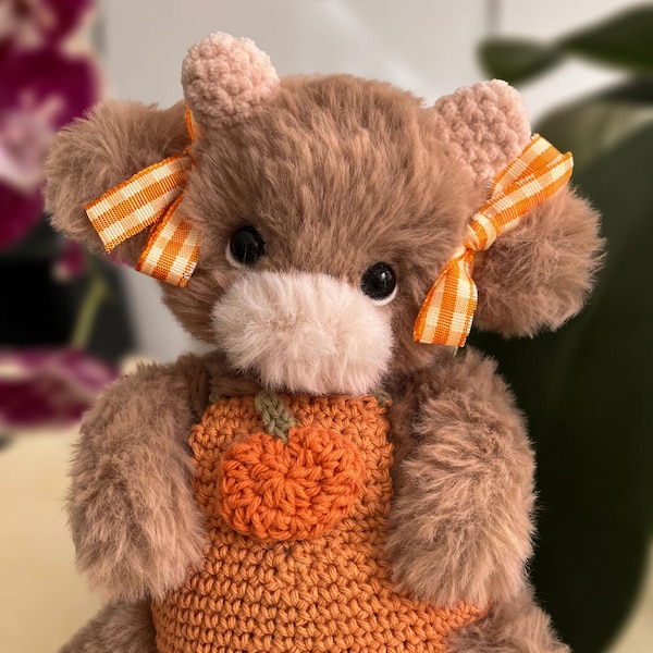 May include: A plush brown teddy bear with fuzzy fur, featuring a light pink muzzle and small rounded ears. The bear wears two orange and white gingham bows on its ears and a crocheted orange pumpkin patch on its chest over a textured body.