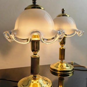 Könnte beinhalten: Zwei Vintage-Tischlampen mit Messingfuß und -schaft, gekrönt von einem weißen, gerüschten Glasschirm. Die Lampen haben ein klassisches Design mit rundem Sockel und dekorativer Spitze. Die Lampen stehen auf einer schwarzen Oberfläche.