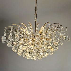Könnte beinhalten: Ein goldfarbener Kronleuchter mit zahlreichen transparenten Kristallkugel-Akzenten. Die Leuchte hat eine Kette zum Aufhängen und gebogene Arme, die die Kristallkugeln halten. Das Design ist aufwendig und elegant, geeignet für die Innenbeleuchtung.