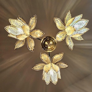 Könnte beinhalten: Eine Deckenleuchte mit drei blütenförmigen Lampen. Jede Lampe hat mehrere Blütenblätter in Gelb- und Weißtönen mit goldenen Verzierungen und dekorativen Mustern. Die zentrale Halterung ist goldfarben, und das Licht wirft einen warmen Schein.