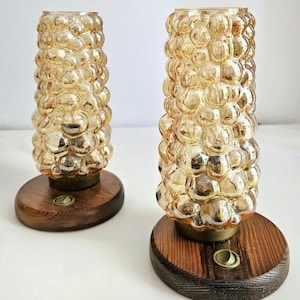 Könnte beinhalten: Zwei zylindrische Tischlampen mit bernsteinfarbenen Glasschirmen mit einer texturierten, blasenartigen Oberfläche. Jede Lampe ist auf einem runden, dunkel gebeizten Holzsockel montiert, in den ein kleiner, runder messingfarbener Druckschalter eingelassen ist.