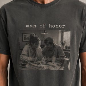 K&ouml;nnte beinhalten: Dunkelgraues T-Shirt mit dem Text "man of honor" &uuml;ber einem Schwarz-Wei&szlig;-Bild von zwei Personen, die ein Puzzle bearbeiten. Das Bild zeigt einen Tisch, eine Tasse und ein Fenster.