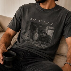 K&ouml;nnte beinhalten: Dunkelgraues T-Shirt mit dem Text "man of honor" &uuml;ber einem Schwarz-Wei&szlig;-Foto von zwei Personen. Das T-Shirt wird von einer Person getragen, die auf einem beigen Sofa sitzt und eine schwarze Tasse h&auml;lt. Das Shirt hat einen Vintage-Look.