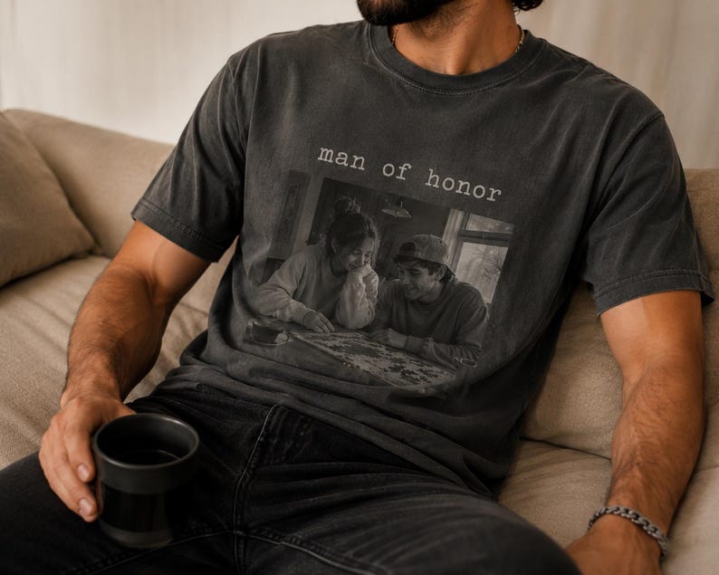 K&ouml;nnte beinhalten: Dunkelgraues T-Shirt mit dem Text "man of honor" &uuml;ber einem Schwarz-Wei&szlig;-Foto von zwei Personen. Das T-Shirt wird von einer Person getragen, die auf einem beigen Sofa sitzt und eine schwarze Tasse h&auml;lt. Das Shirt hat einen Vintage-Look.