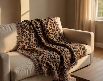 May include: A woven throw blanket featuring a brown, tan, and black leopard print pattern. The rectangular textile has a textured surface and is finished with short, frayed fringe detailing along the edges, creating a soft and tactile appearance.