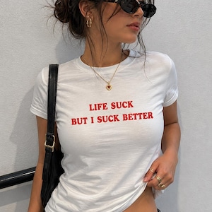 Könnte beinhalten: Weißes T-Shirt mit dem roten Text "LIFE SUCK BUT I SUCK BETTER". Das Shirt hat kurze Ärmel und ist figurbetont. Die Person trägt eine Sonnenbrille, Goldschmuck und eine dunkelblaue Jeans. Eine schwarze Umhängetasche ist ebenfalls zu sehen.