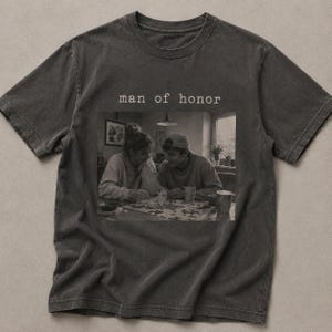 K&ouml;nnte beinhalten: Dunkelgraues T-Shirt mit dem Text "man of honor" &uuml;ber einem Schwarz-Wei&szlig;-Bild von zwei Personen, die ein Puzzle machen. Das Bild ist mittig auf dem Shirt platziert.