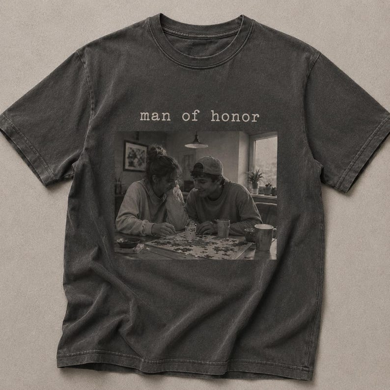 K&ouml;nnte beinhalten: Dunkelgraues T-Shirt mit dem Text "man of honor" &uuml;ber einem Schwarz-Wei&szlig;-Bild von zwei Personen, die ein Puzzle machen. Das Bild ist mittig auf dem Shirt platziert.