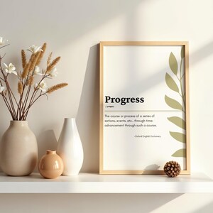 Puede incluir: Impresión enmarcada con la palabra "Progress" y su definición del Diccionario Oxford English. La impresión está en un estante blanco con jarrones y un pequeño objeto redondo y marrón.