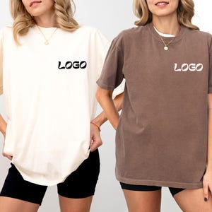 Puede incluir: Dos camisetas de manga corta, una marfil y otra espresso, cada una con la palabra "LOGO" impresa en la parte delantera. Las camisetas se usan con pantalones cortos negros. La camiseta marfil está a la izquierda y la camiseta espresso a la derecha.