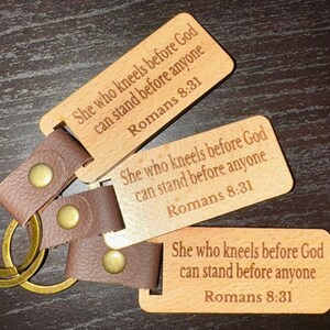 Könnte beinhalten: Holz-Schlüsselanhänger mit braunen Lederriemen und goldfarbener Hardware. Jeder Schlüsselanhänger trägt den eingravierten Text: "She who kneels before God can stand before anyone Romans 8:31".