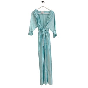 May include: A light blue floor-length robe featuring a V-neckline with ruffled trim, three-quarter length sleeves with ruffled cuffs, and a matching fabric tie belt at the waist. The fabric has a subtle tonal floral pattern throughout the garment.