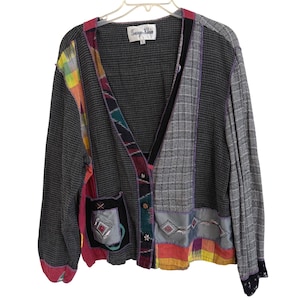 Op de afbeelding: Een grijze cardigan met een patchwork ontwerp. De trui heeft een V-hals, lange mouwen en een knoopsluiting. Kleurrijke stoffen patches zijn op de voorkant en mouwen genaaid, met een label met de tekst "George Klein".