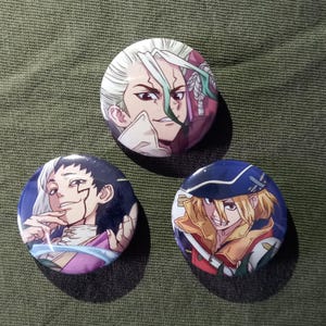 Può includere: Tre spille rotonde con personaggi anime. Le spille presentano illustrazioni colorate di personaggi con espressioni facciali e abiti dettagliati. Lo sfondo è verde oliva.