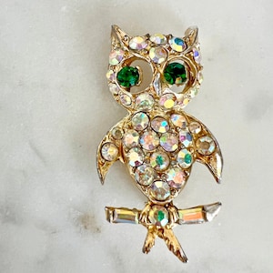 May include: A gold-toned owl brooch featuring iridescent, multi-colored rhinestones covering the body and head. The owl has two prominent, round, deep green rhinestone eyes and is perched on a horizontal branch accented with clear crystals.