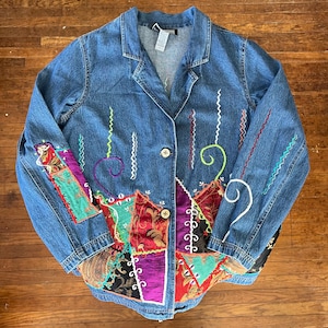May include: A blue denim jacket with colorful patchwork and embroidered accents. The jacket features a button-up front, a collar, and decorative stitching in various colors, including red, green, and white. Patchwork includes paisley and floral patterns.