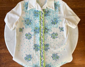 May include: A white short-sleeved button-up shirt with a floral pattern in shades of blue, green, and white. The shirt has a collar and a button-up front.