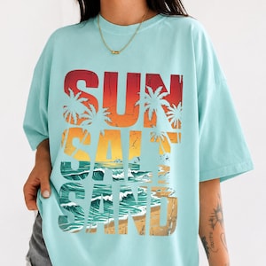 May include: A light blue t-shirt with the words "SUN SALT SAND" in a gradient design of red, orange, yellow, and brown. The design includes palm trees, ocean waves, and a beach scene.
