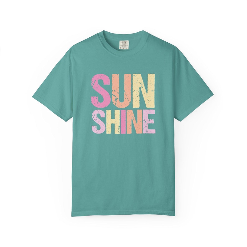 Teal t-shirt with 'SUNSHINE' graphic in distressed lettering, 'SUN' in pink and orange, 'SHINE' in pink, yellow, and orange