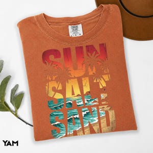 May include: An orange Comfort Colors t-shirt with a graphic that reads "SUN SALT SAND" in a beach-themed design. The design features palm trees, ocean waves, and a sunset color scheme. A brown hat is partially visible.