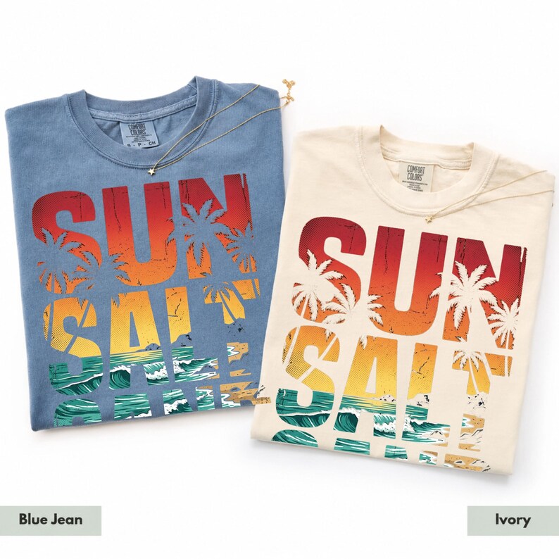 May include: Two t-shirts, one blue and one ivory, with the words "SUN SALT" in a gradient of red, orange, and yellow, with palm trees and ocean waves. The blue shirt is labeled "Blue Jean" and the ivory shirt is labeled "Ivory".