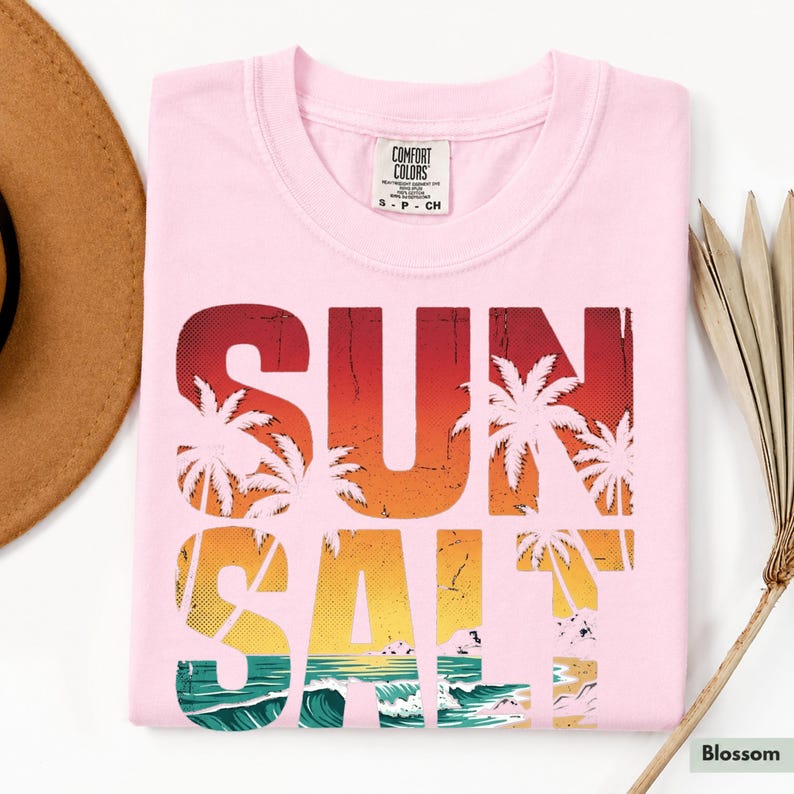 May include: A light pink t-shirt with the words "SUN SALT" in a distressed, colorful design featuring palm trees, a sunset, and ocean waves. The shirt has a crew neck and a small tag at the neckline.
