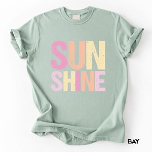 SUNSHINE' printed on light green t-shirt with distressed 'SUN' in pink and orange, 'SHINE' in pink, yellow, and orange
